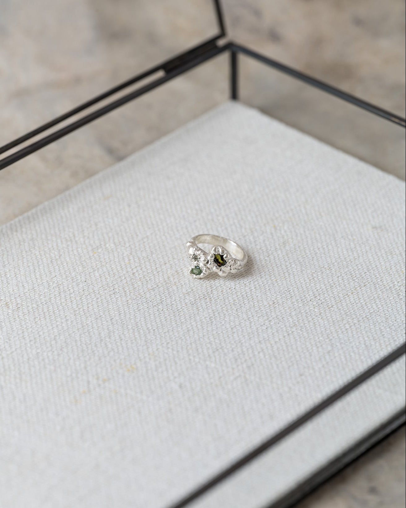 Silver ring with a green gemstone on a gray cushion in a glass display case.