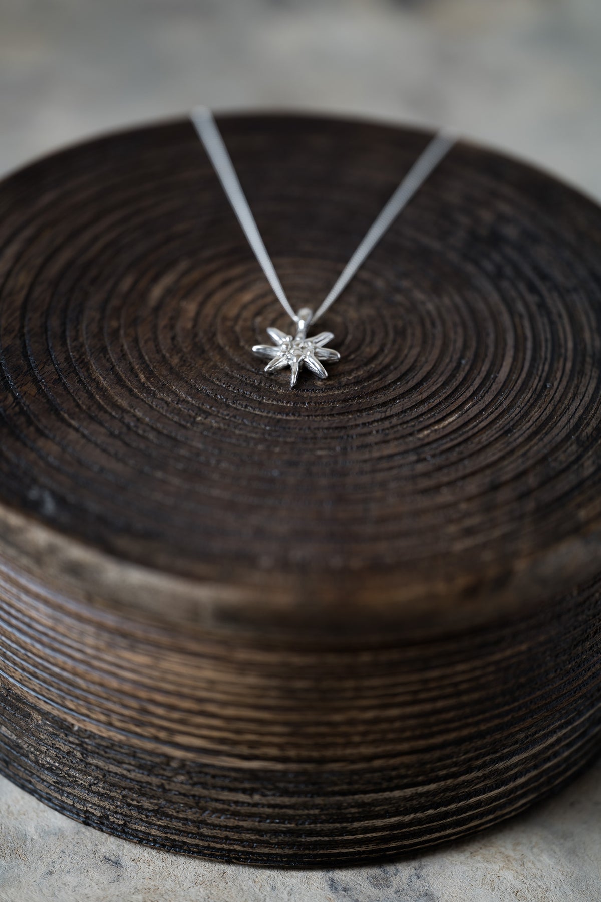 A silver snowflake pendant necklace on a delicate silver chain, displayed on a dark brown wooden slice prop against a soft gray background.