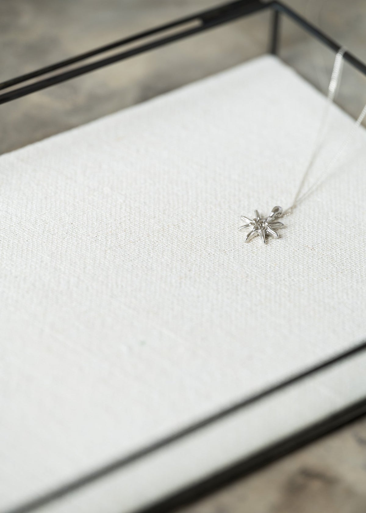 A delicate silver snowflake pendant necklace resting on a textured white fabric surface inside a black glass jewelry display box.
