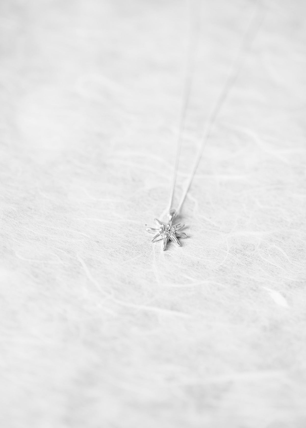 A delicate silver necklace with a small starburst or snowflake pendant, resting on a textured white surface.