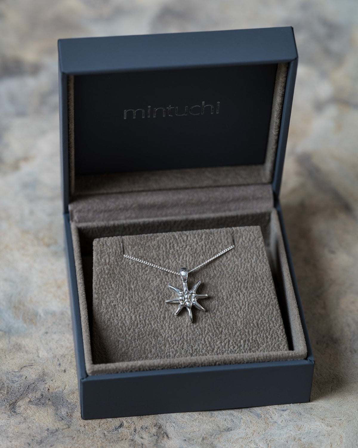 A silver starburst pendant necklace displayed inside an open dark gray mintuchi jewelry box on a textured stone surface.