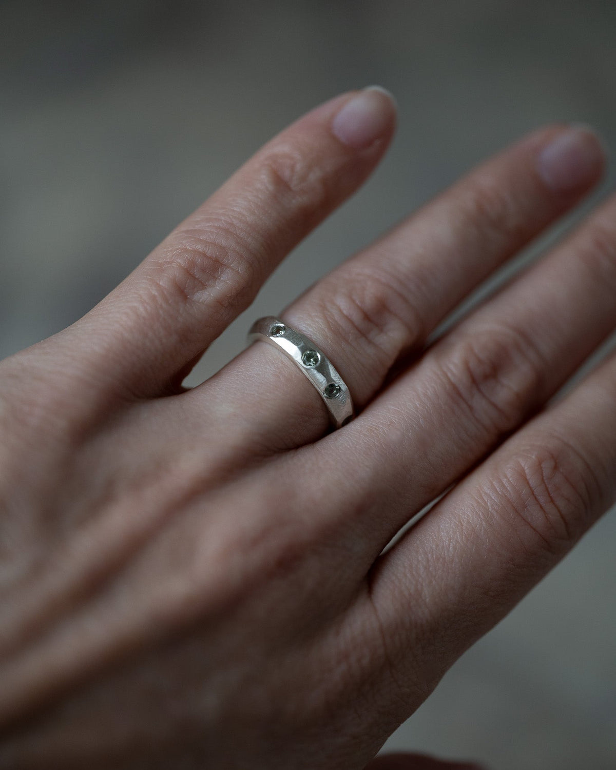 A close-up of a hand wearing a simple polished silver band ring, shown against a soft, out-of-focus gray background.