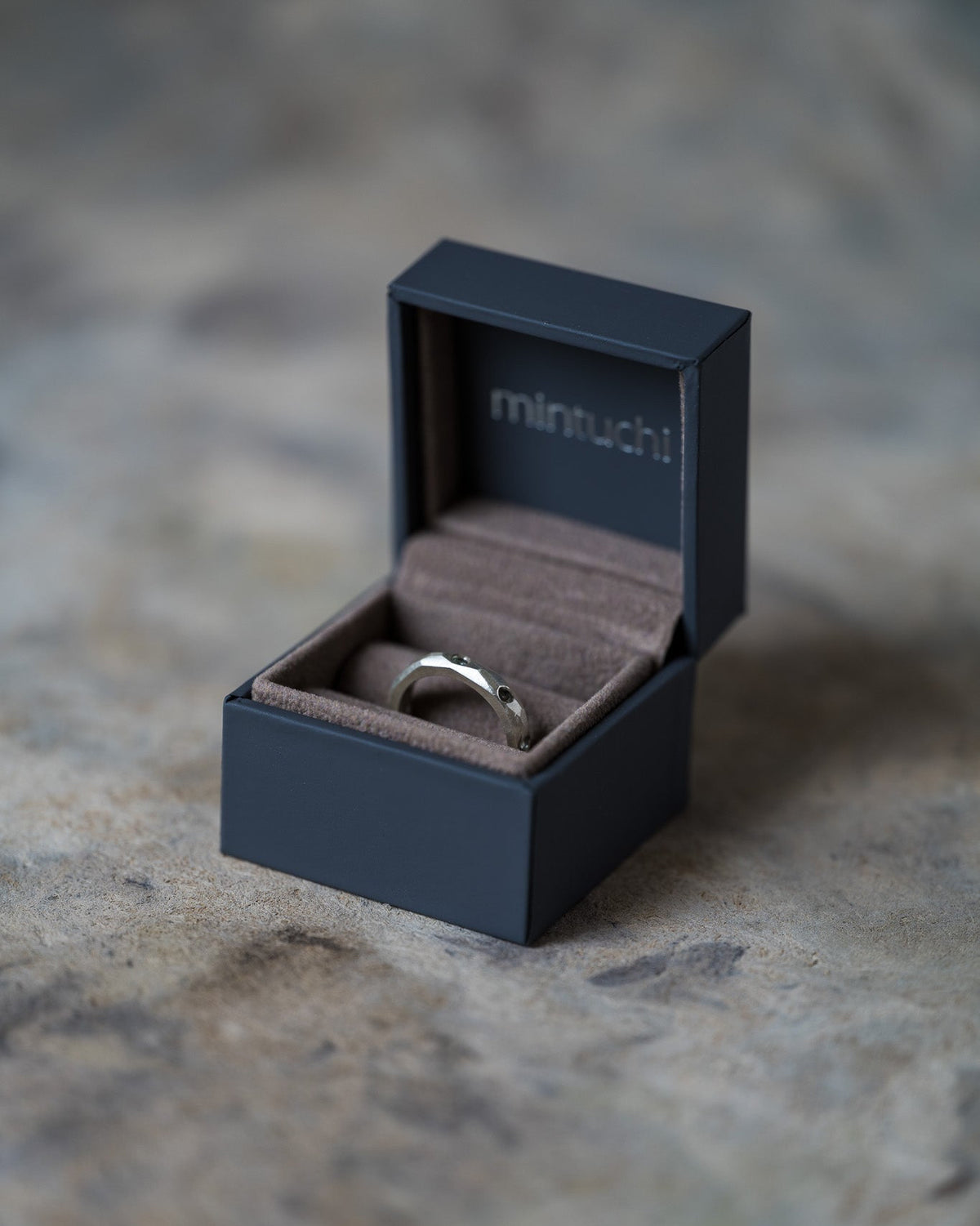 A silver band ring with a small black stone sits inside an open dark navy Mintuchi jewelry box on a textured stone surface.