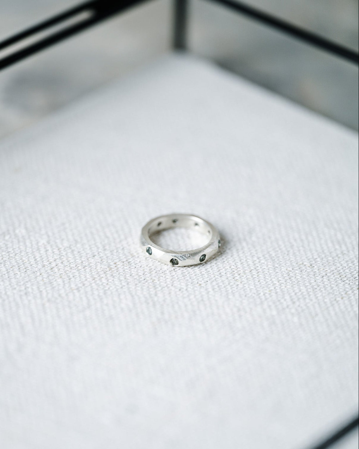 A silver band ring with small dark round stones, resting on a textured white fabric surface inside a glass display case.