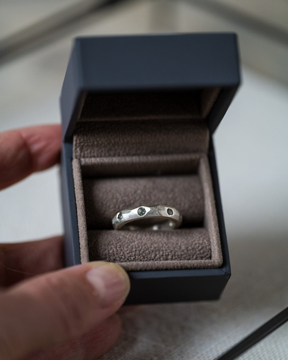 A silver ring with three small green gemstones set into a dark gray velvet ring box, held by a hand.