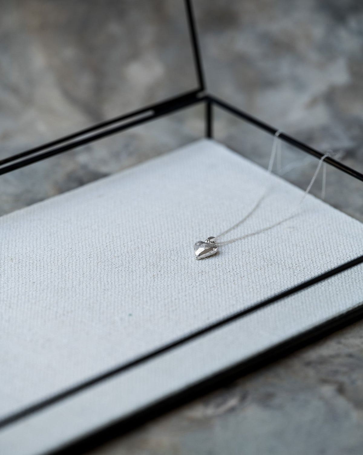 A delicate silver necklace with a small heart-shaped pendant resting on a white textured display cushion inside a black glass jewelry box, set against a gray stone background.