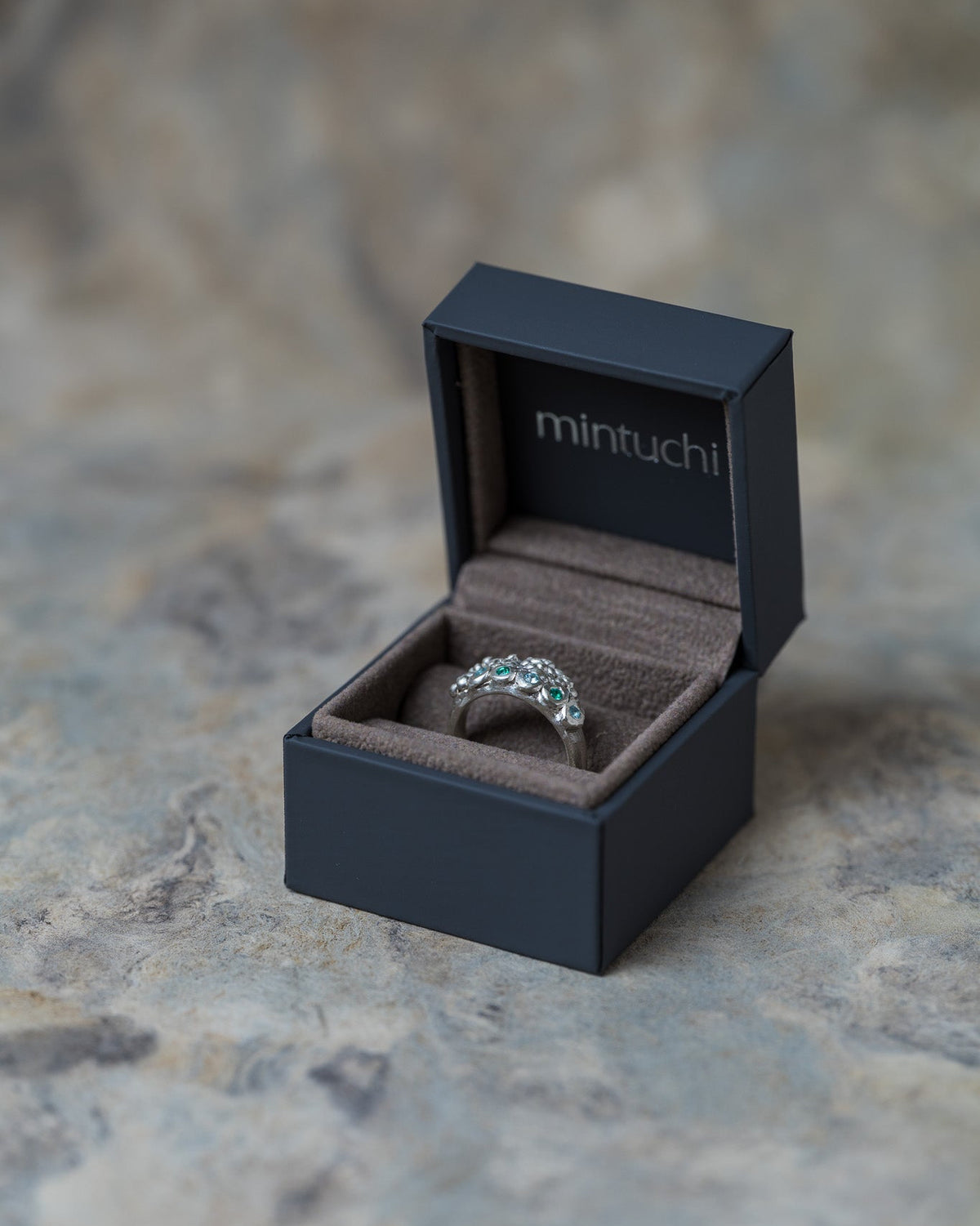 A silver ring with a floral cluster setting sits inside an open black Mintuchi jewelry box on a textured stone surface.