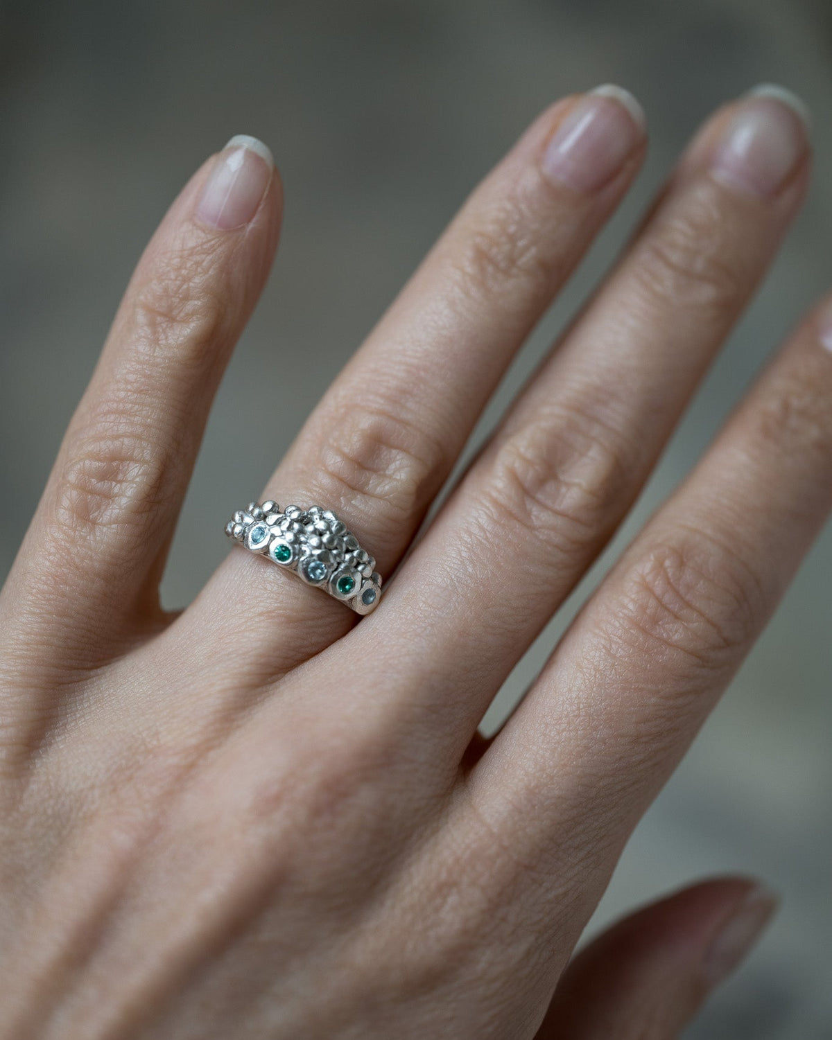 A close-up of a hand wearing a silver band ring decorated with small round-cut clear stones and a row of tiny green gem accents.