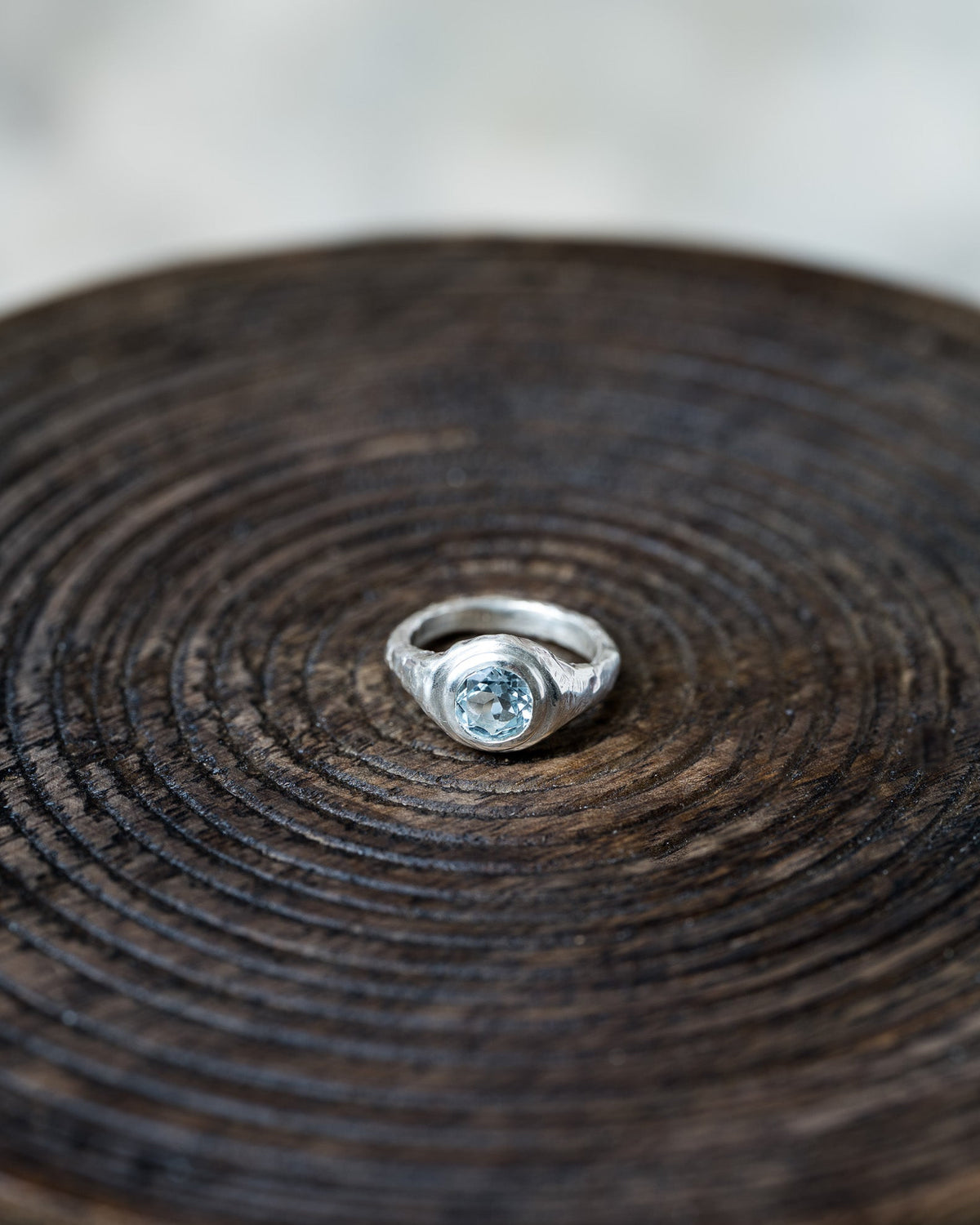 A silver ring with a round light blue gemstone, displayed on a dark brown wooden slice background.