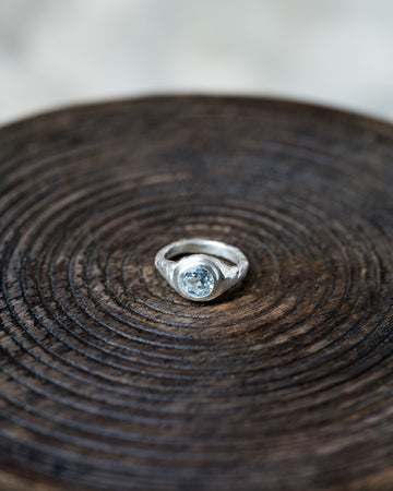 A silver ring with a round light blue gemstone, displayed on a dark brown wooden slice background.