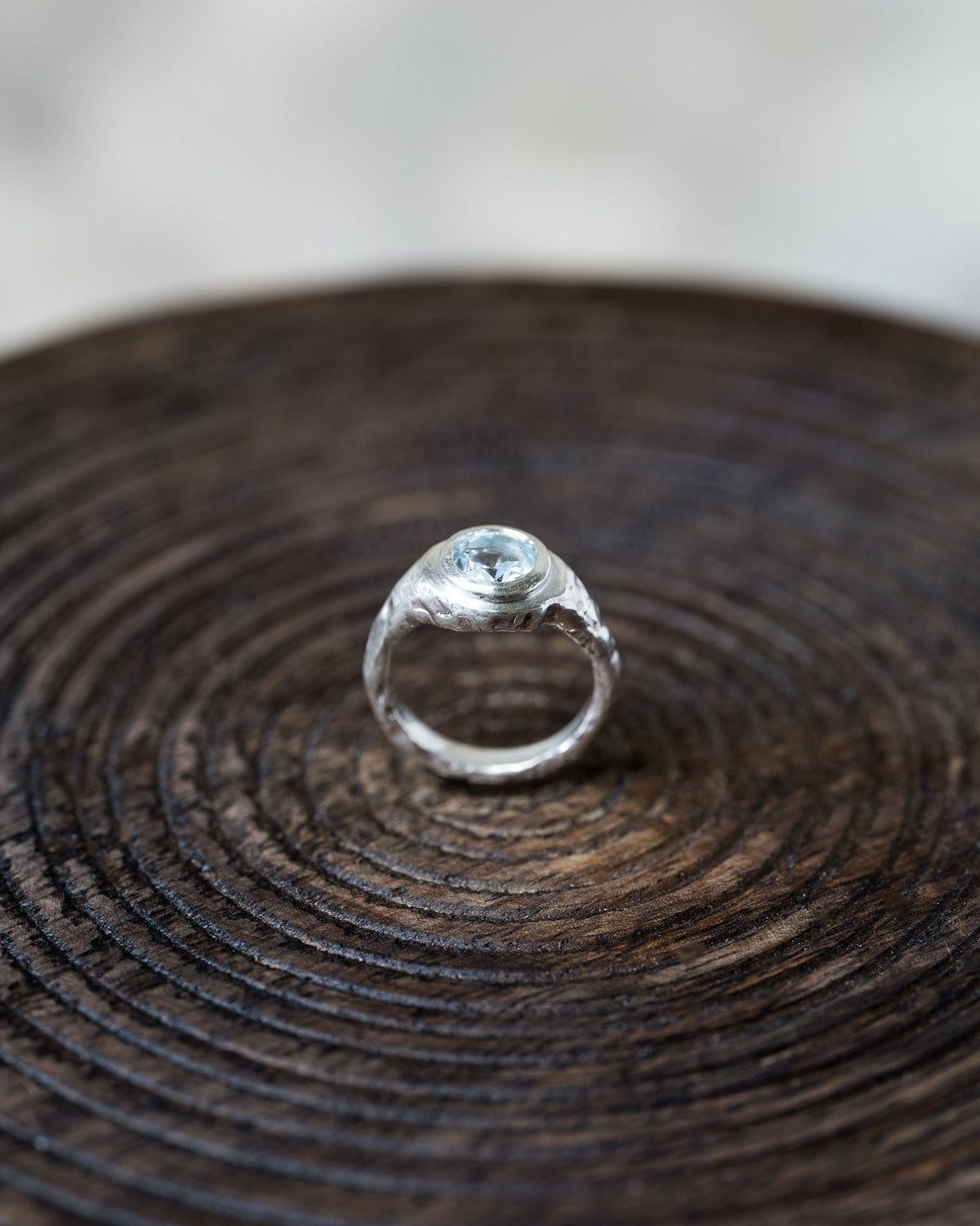 A silver ring with a textured, organic-shaped setting and a small round clear gemstone, displayed on a dark wood slice background.