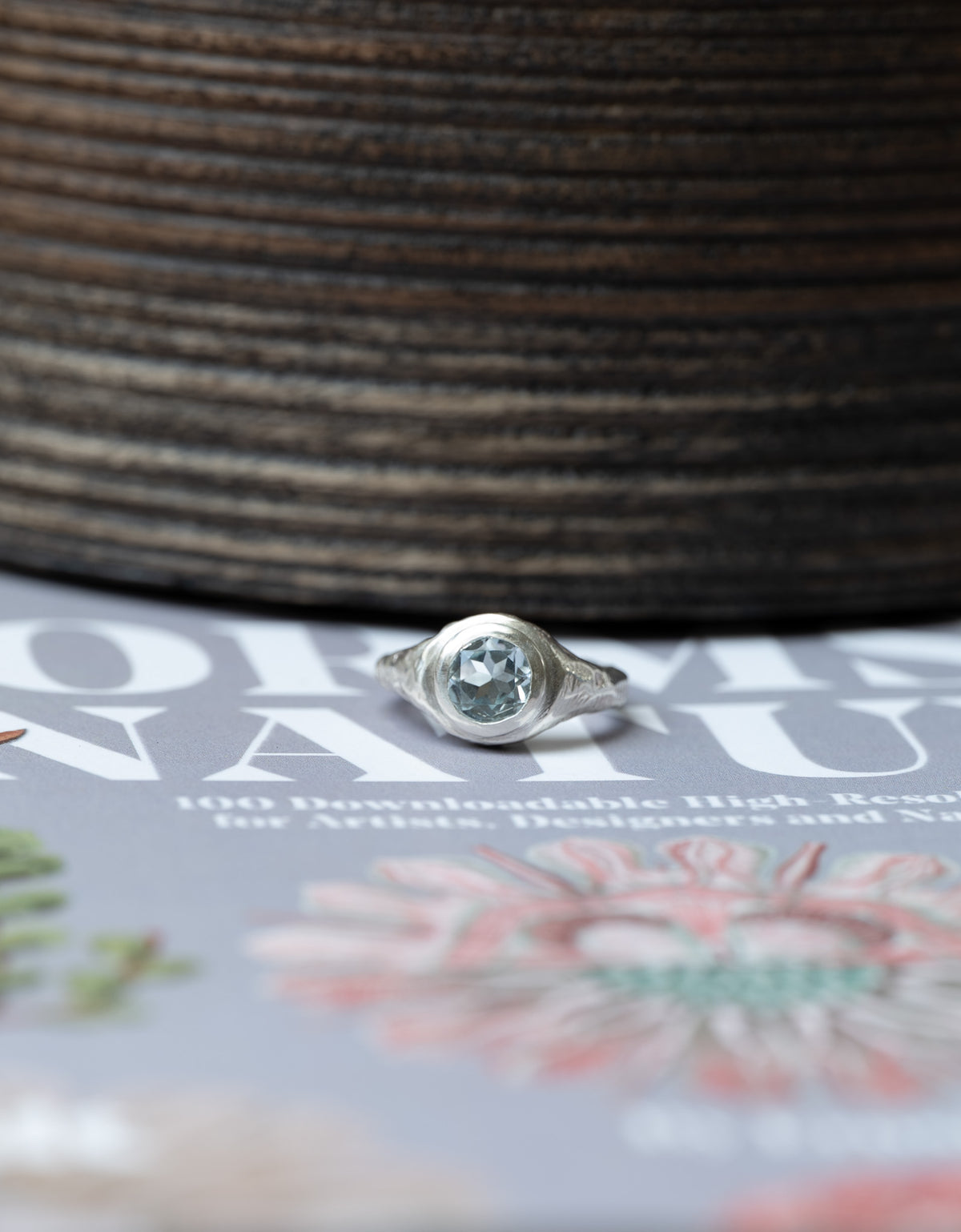 A silver ring with a round, bezel-set light blue gemstone resting on a printed magazine page, with a dark brown textured wooden surface in the background.