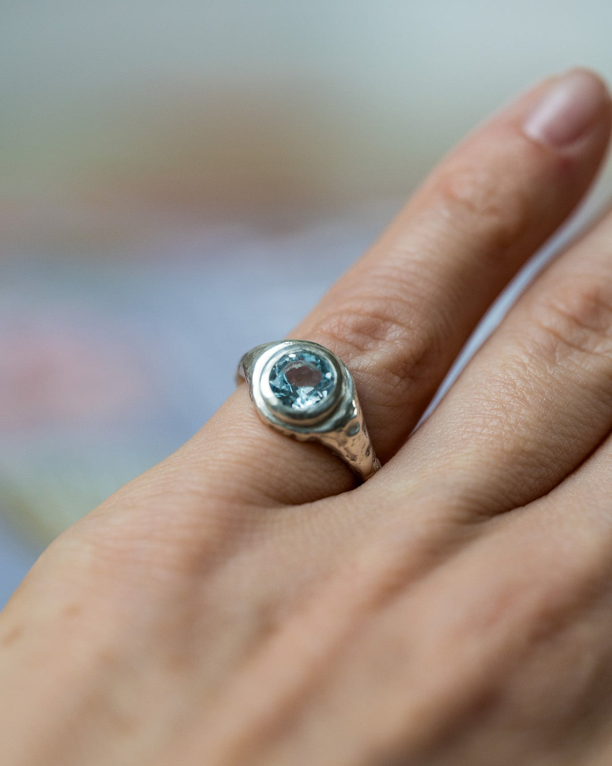 A silver ring with a round light blue gemstone set on a person's ring finger, shown in a close-up shot with a soft, blurred background.