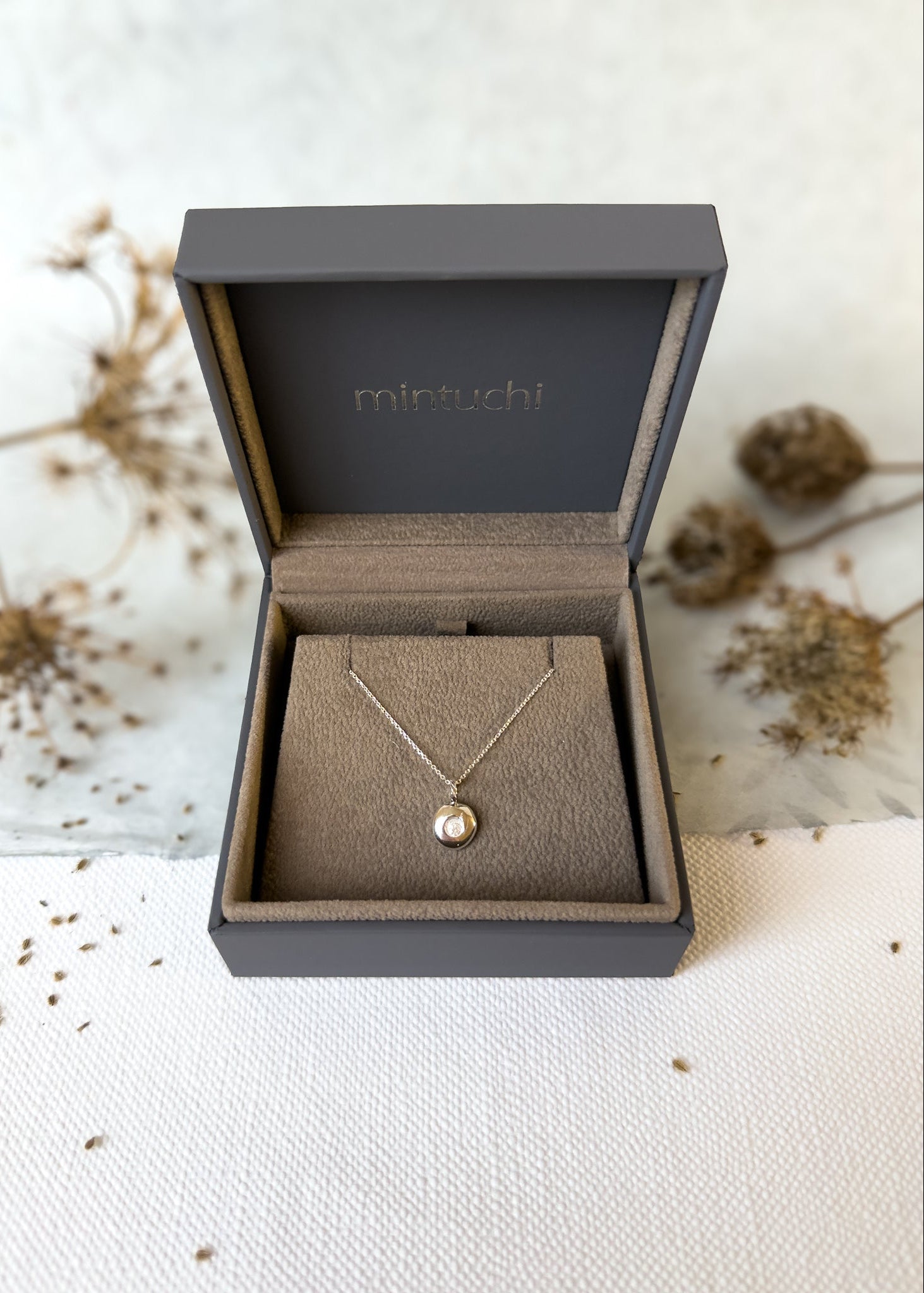Necklace in a 'mintuchi' branded box on a textured surface with dried flowers.