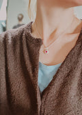 Close-up of a person wearing a necklace with a red pendant, wearing a brown cardigan and light blue top.