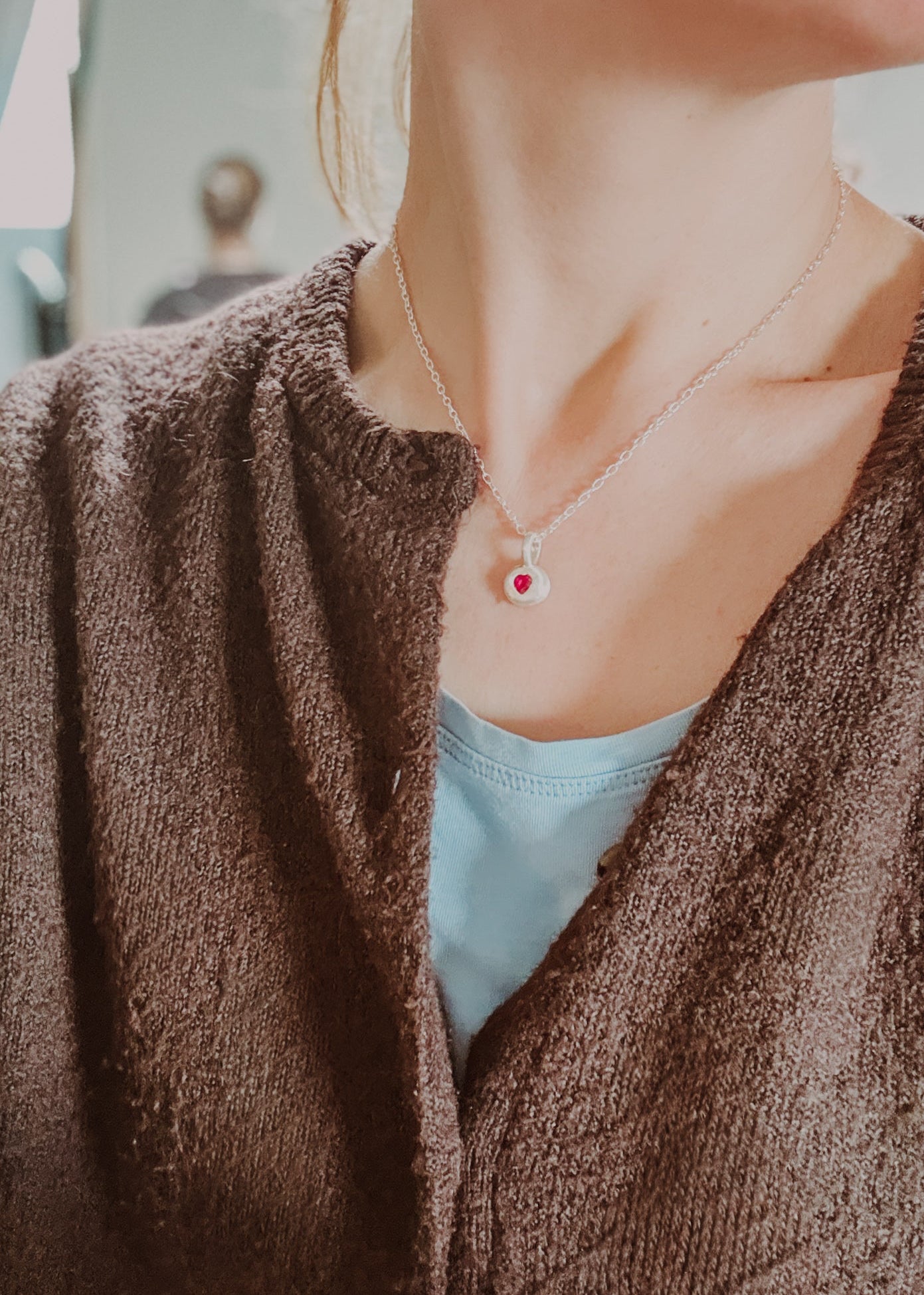 Close-up of a person wearing a necklace with a red pendant, wearing a brown cardigan and light blue top.