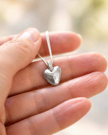 A silver heart pendant necklace with a textured, faceted finish hanging from a delicate silver chain, held in a person's hand against a soft, blurred background.
