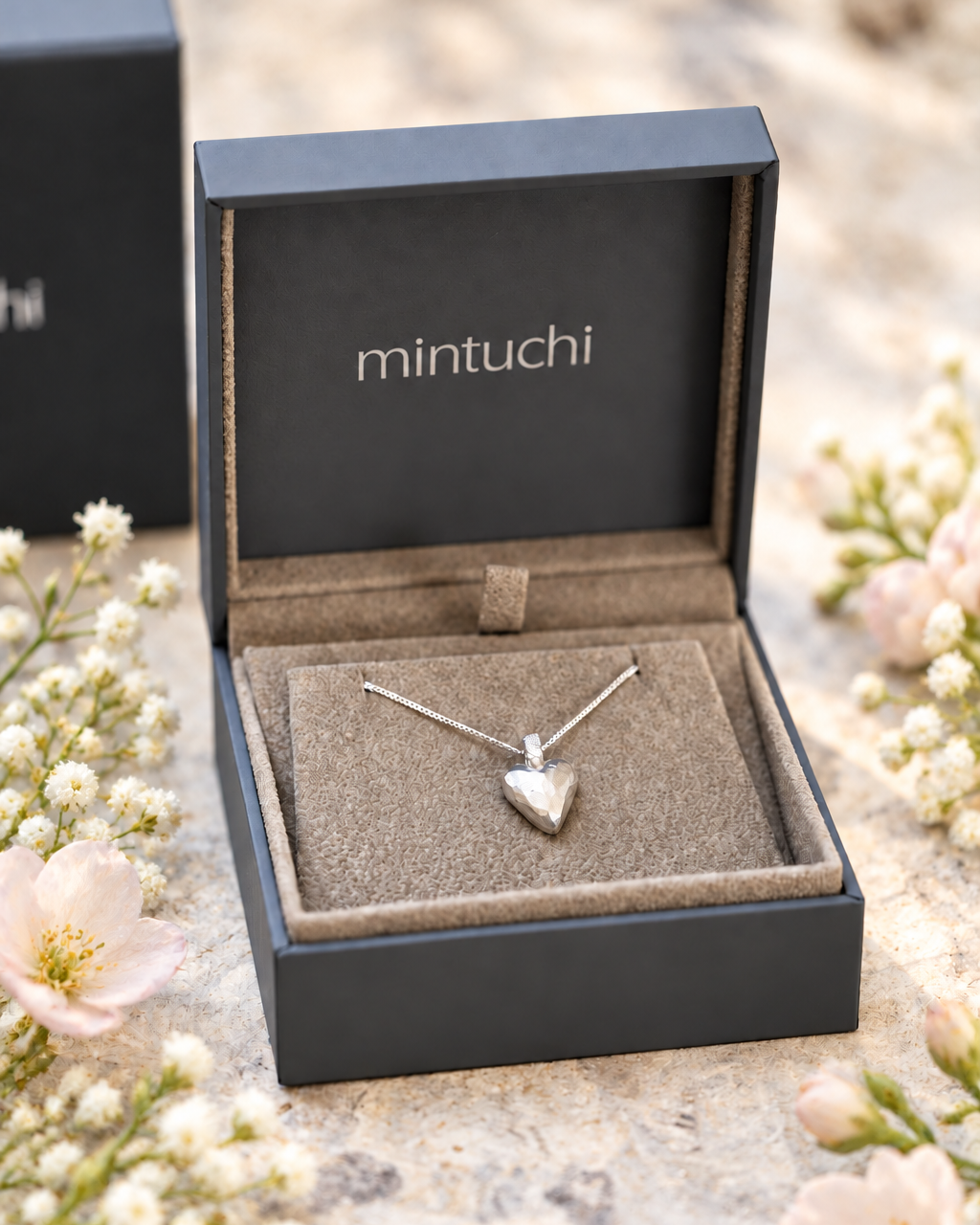 A silver heart pendant necklace on a delicate chain, presented in an open black Mintuchi jewelry box with a beige velvet insert, surrounded by soft pink flowers on a textured stone surface.