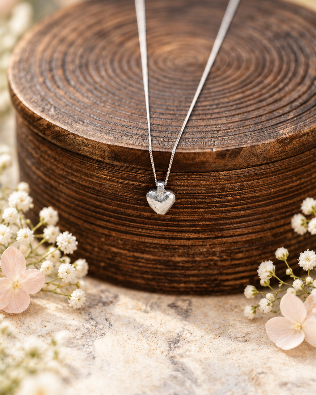 A silver heart pendant necklace on a delicate chain, displayed on a round wooden slice with small white flowers around it.